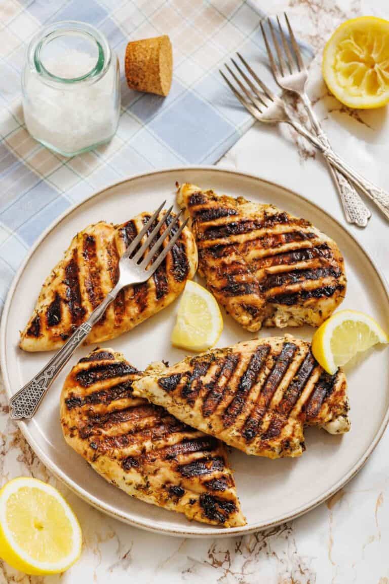 A plate with four pieces of grilled chicken breast, grill marks visible, garnished with lemon wedges; two forks, a jar of salt, and halved lemons are nearby on a table.