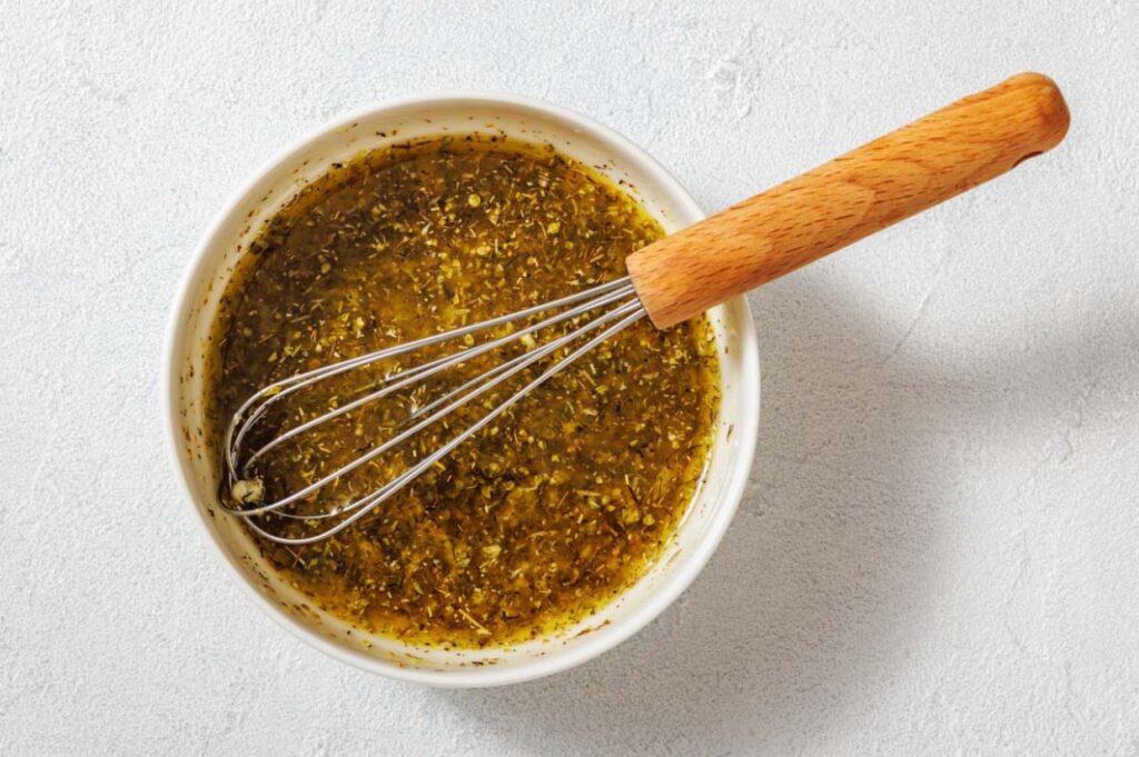 A bowl of vinaigrette dressing with herbs being mixed by a metal whisk on a light-colored surface.