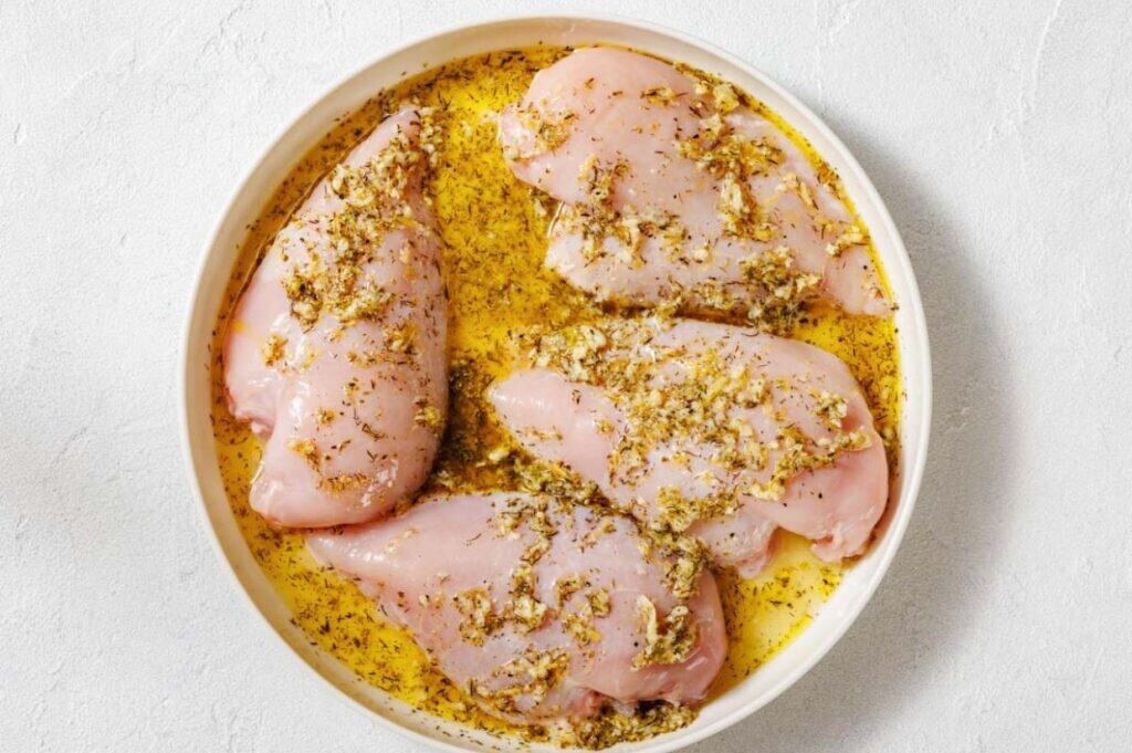 Four raw chicken breasts marinated with herbs and oil are arranged in a round white dish on a white surface.