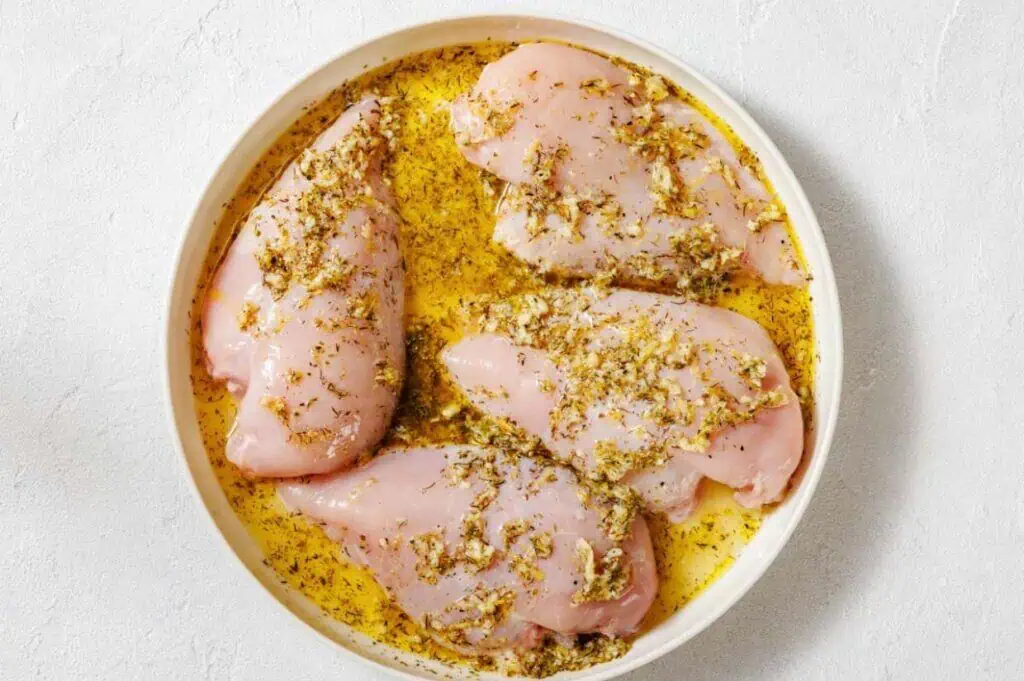 Four raw chicken breasts marinated with herbs and oil are arranged in a round white dish on a white surface.