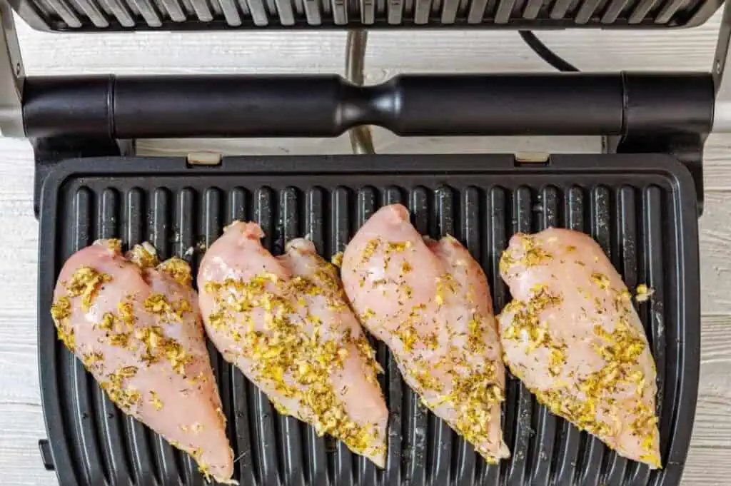 Four seasoned raw chicken breasts placed on the grill plates of an electric indoor grill, ready to be cooked.