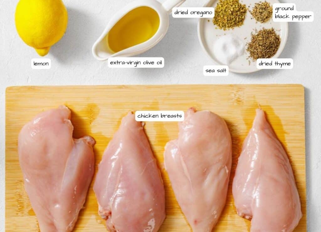 Four raw chicken breasts on a wooden board with lemon, extra-virgin olive oil, sea salt, dried thyme, dried oregano, and ground black pepper arranged nearby.
