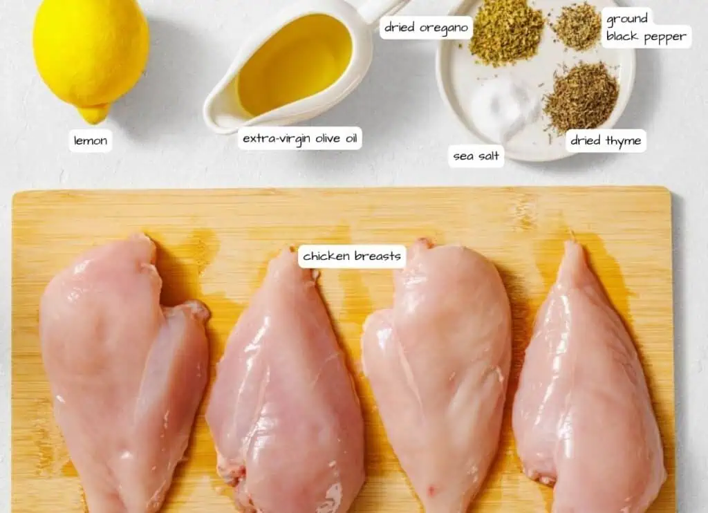 Four raw chicken breasts on a wooden board with lemon, extra-virgin olive oil, sea salt, dried thyme, dried oregano, and ground black pepper arranged nearby.