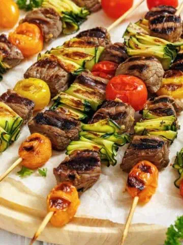 Grilled skewers with beef, zucchini slices, and cherry tomatoes are arranged on a wooden platter, garnished with parsley.