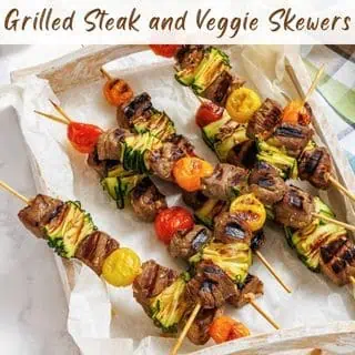 Wooden tray with grilled steak and vegetable skewers, featuring pieces of steak, zucchini, and cherry tomatoes, arranged on parchment paper. Text at top reads: "Grilled Steak and Veggie Skewers.