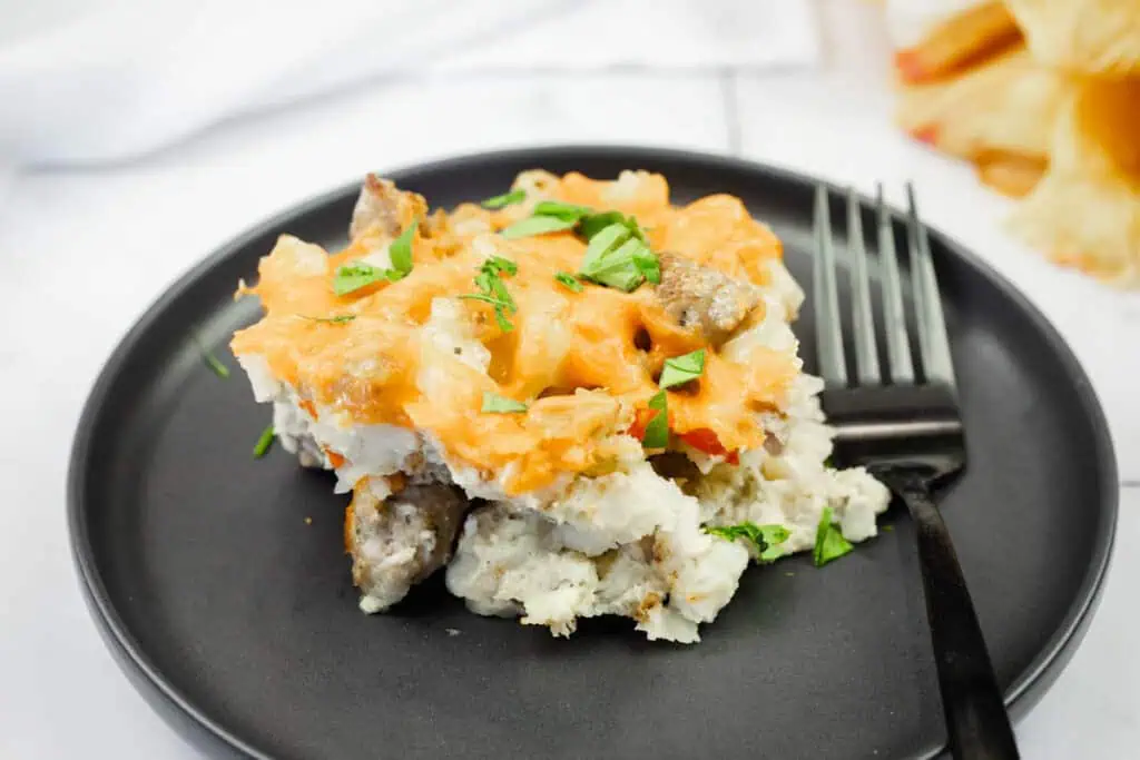 A serving of cheesy breakfast casserole with sausage and herbs on a black plate with a fork.