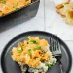 A black plate with a serving of cheesy casserole containing meat and herbs, placed next to a fork, with a baking dish of casserole and flowers in the background.