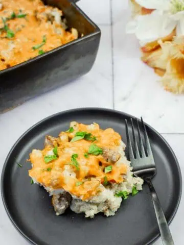 A black plate with a serving of cheesy casserole containing meat and herbs, placed next to a fork, with a baking dish of casserole and flowers in the background.
