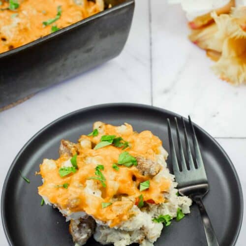 A black plate with a serving of cheesy casserole containing meat and herbs, placed next to a fork, with a baking dish of casserole and flowers in the background.