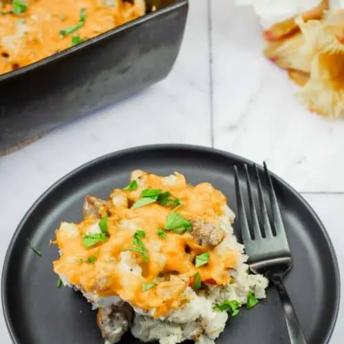 A black plate with a serving of cheesy casserole containing meat and herbs, placed next to a fork, with a baking dish of casserole and flowers in the background.