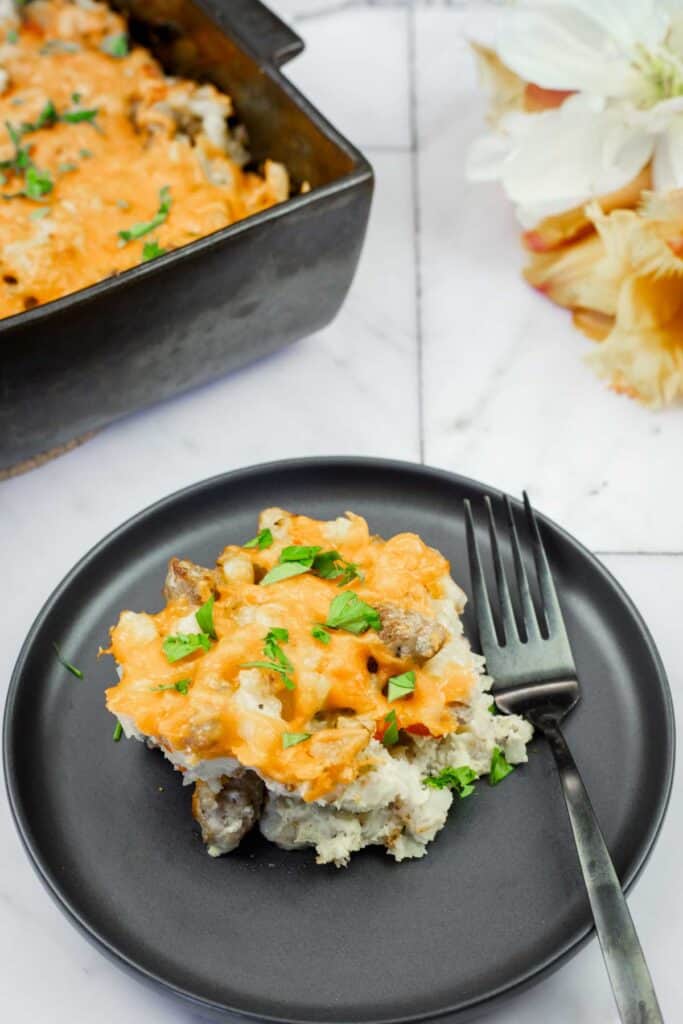 A black plate with a serving of cheesy casserole containing meat and herbs, placed next to a fork, with a baking dish of casserole and flowers in the background.