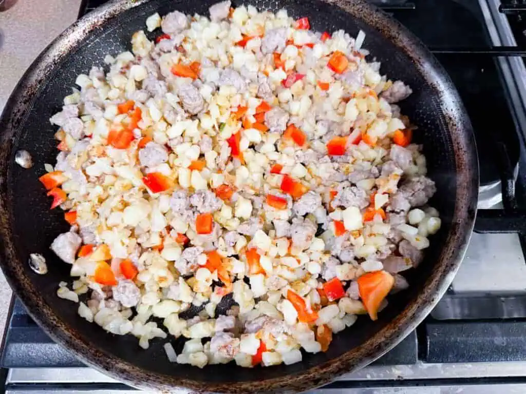 A frying pan on a stove contains diced onions, red bell peppers, and sausage being sautéed.