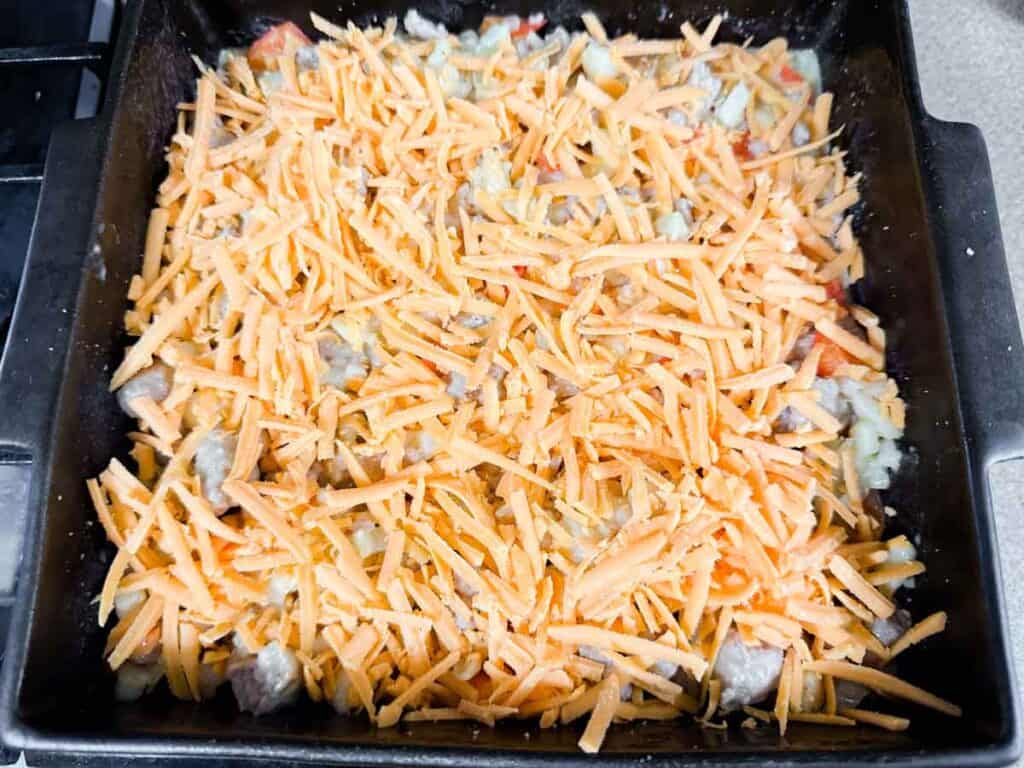 A baking dish filled with a mixture of chopped vegetables and meat, topped with a generous layer of shredded cheddar cheese.
