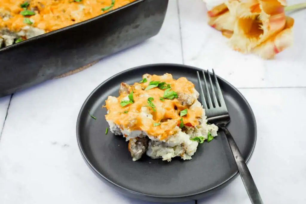 A serving of cheesy casserole with sausage and herbs on a black plate with a fork, beside a baking dish and seashells on a marble surface.