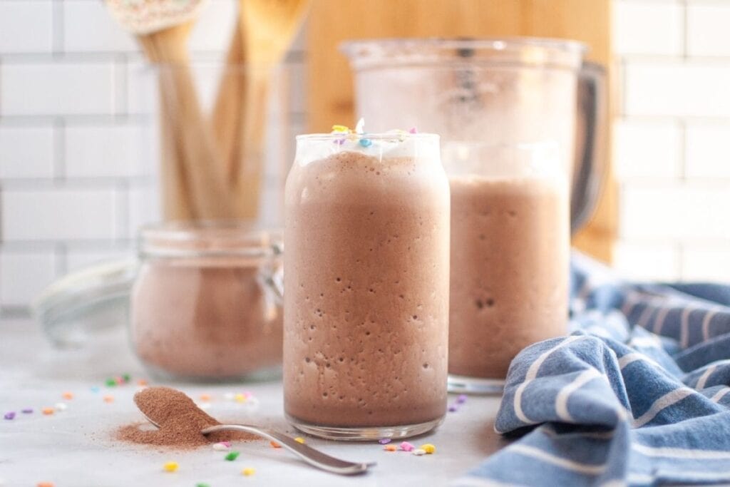 A clear glass filled with frothy brown frozen hot chocolate, topped with a small amount of white whipped cream and colorful sprinkles, on a white countertop with a spoon of cocoa powder.