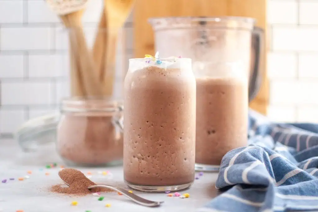 A clear glass filled with frothy brown frozen hot chocolate, topped with a small amount of white whipped cream and colorful sprinkles, on a white countertop with a spoon of cocoa powder.