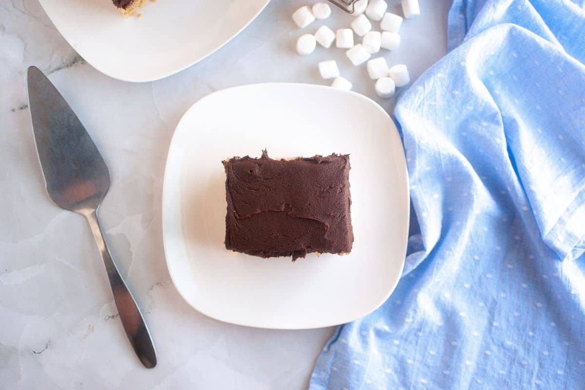 A square slice of Graham Cracker Cake on a white plate, with a cake server, blue cloth, and mini marshmallows nearby.