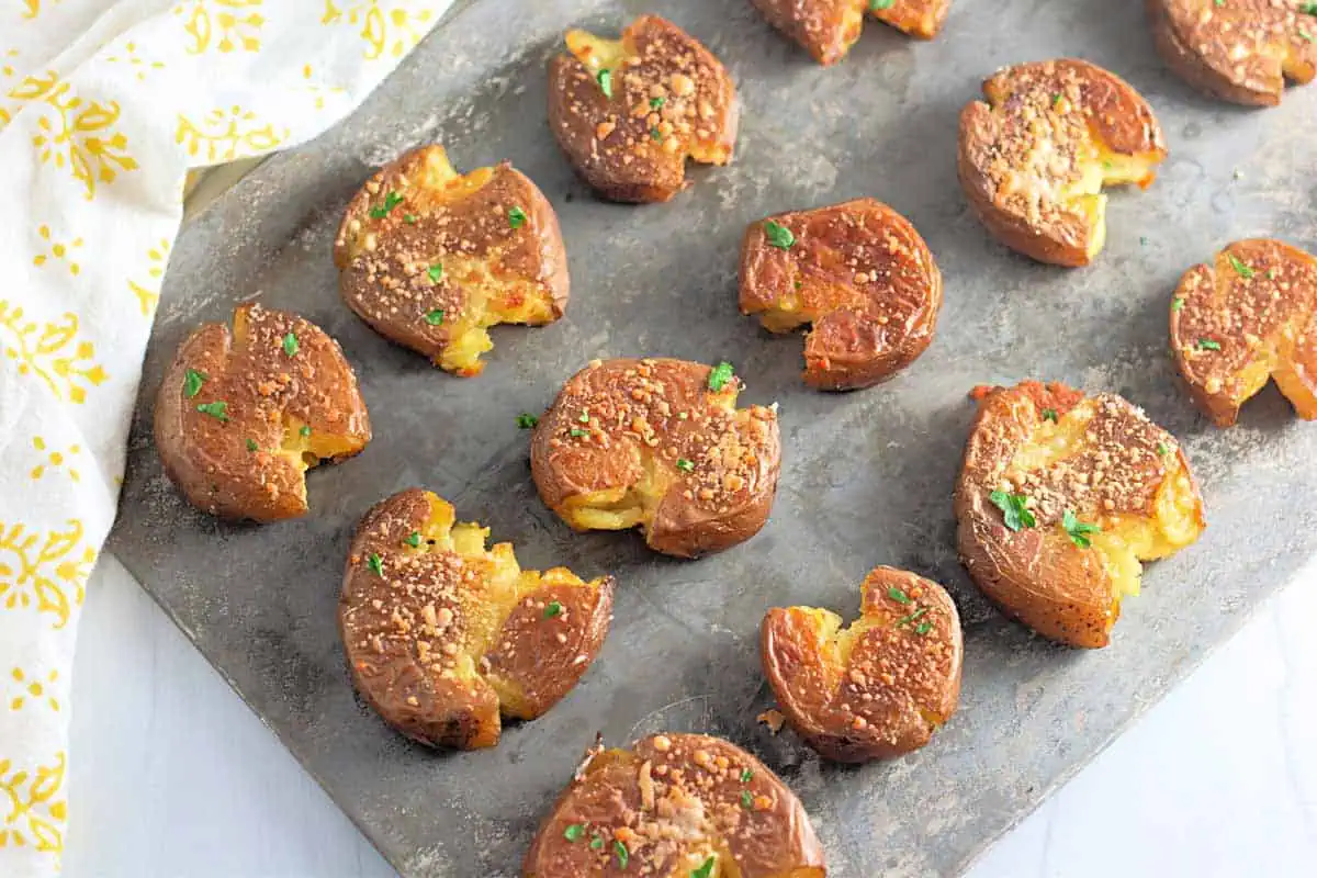 A baking sheet with several smashed roasted potatoes, garnished with herbs and grated cheese, arranged in rows.