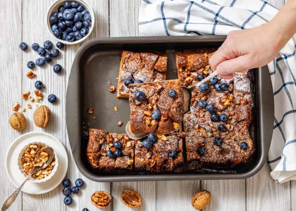 A baking pan with sliced Baked Cinnamon French Toast is shown, with a hand using a spoon to lift one piece.