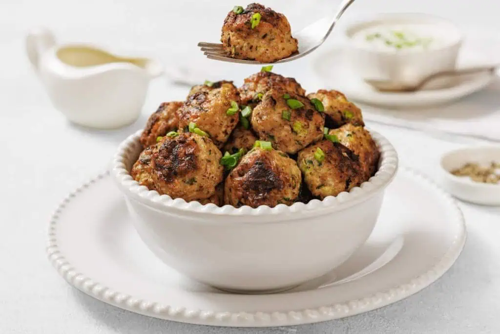 A white bowl filled with Chicken Zucchini Poppers garnished with chopped herbs, with a fork holding one meatball above the bowl