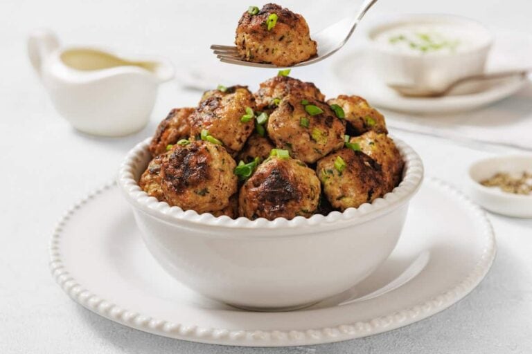 A white bowl filled with Chicken Zucchini Poppers garnished with chopped herbs, with a fork holding one meatball above the bowl