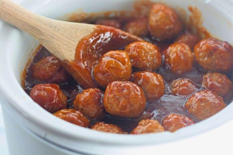 A wooden spoon stirs meatballs coated in a red sauce inside a white slow cooker.