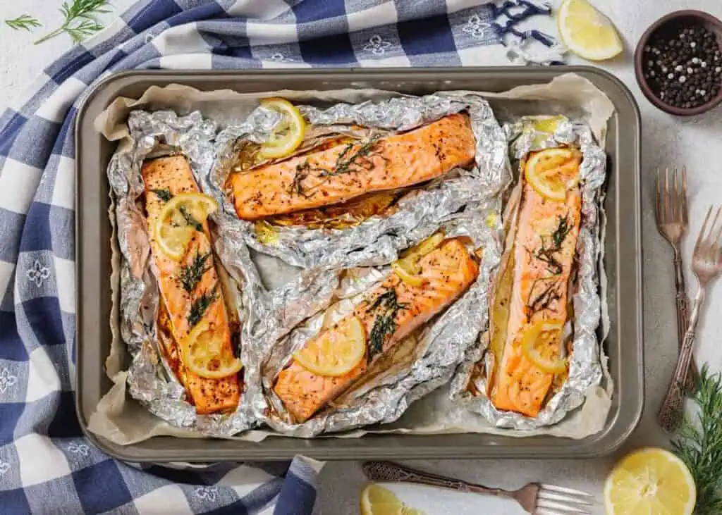 Four individual servings of lemon-dill salmon baked in foil packets, arranged on a baking sheet lined with parchment paper. Lemon slices and fresh dill sprigs are visible on top of the salmon. A blue and white checkered napkin is draped on the upper left, and loose lemons, dill, and a bowl of peppercorns are at the bottom.