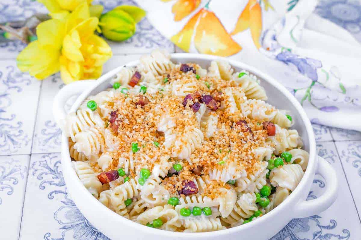 A white bowl filled with Lemon Pasta with Peas & Pancetta, placed on a patterned surface with yellow flowers and a floral napkin nearby.
