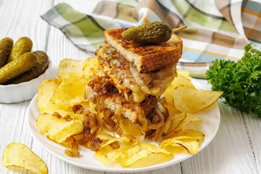 Stacked sandwich with melted cheese and caramelized onions on toasted bread, topped with a pickle, served with potato chips on a white plate.