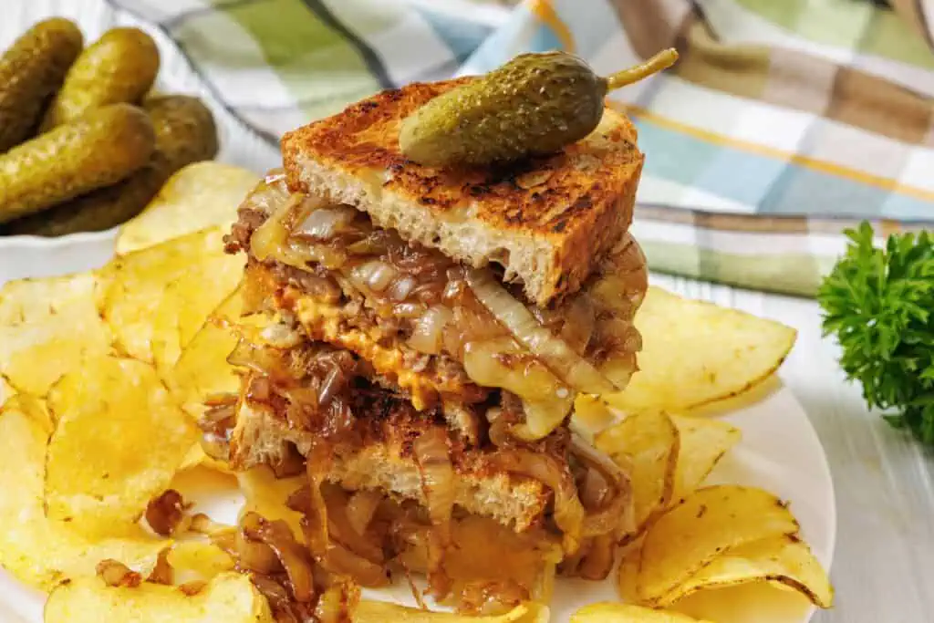 A stacked sandwich with caramelized onions and melted cheese on toasted bread, topped with a pickle, served with potato chips on a white plate.
