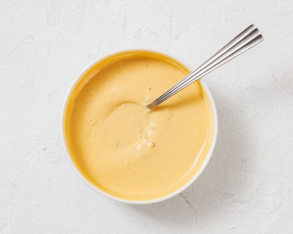 A bowl of creamy yellow cheese sauce with a metal spoon on a light textured surface.
