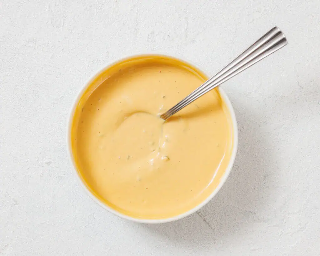 A bowl of creamy yellow cheese sauce with a metal spoon on a light textured surface.