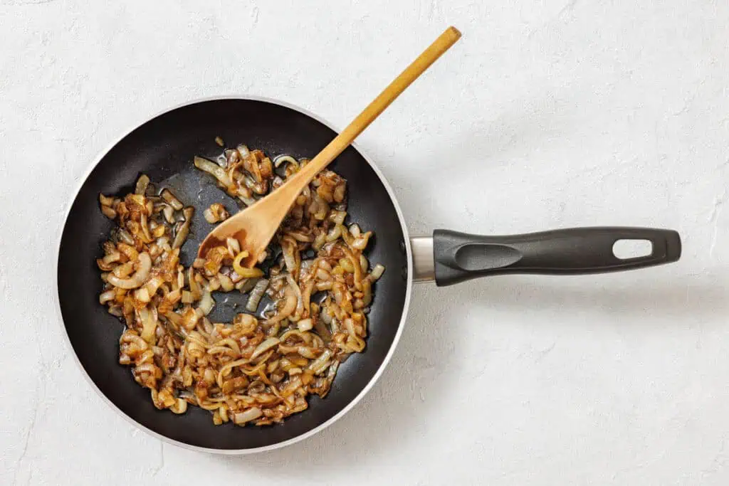 Sliced onions being sautéed in a frying pan with a wooden spoon on a light-colored surface.