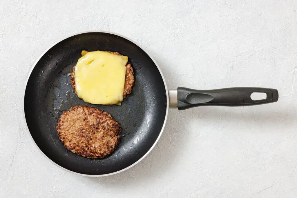 Two cooked burger patties in a frying pan, one topped with melted cheese, on a light-colored surface.