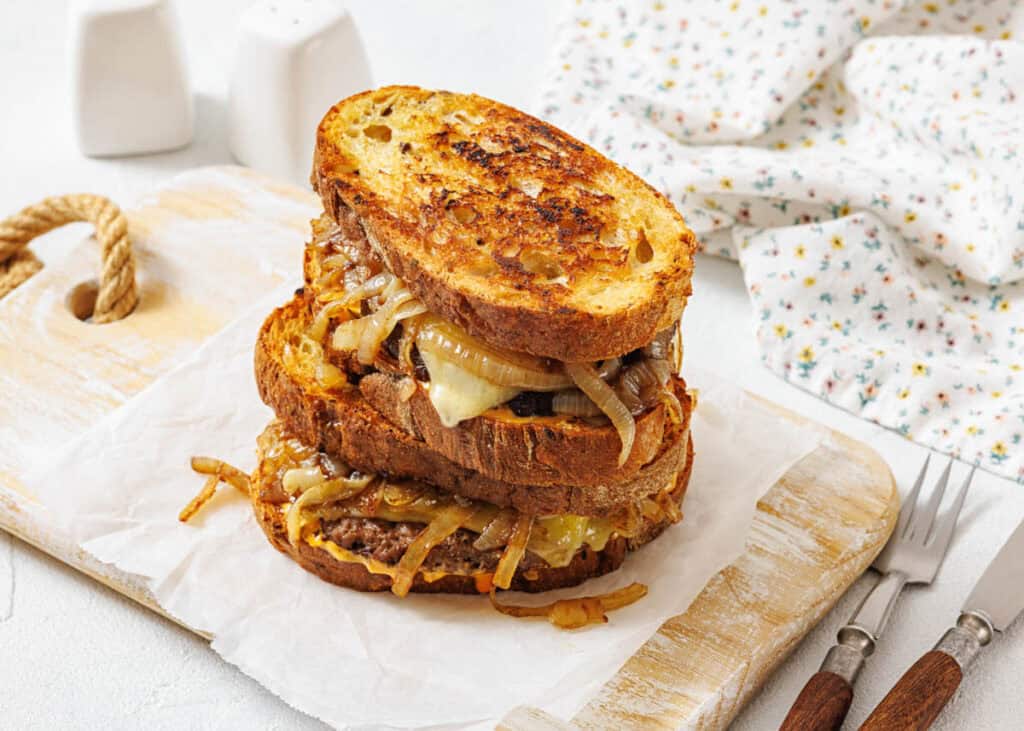 Two patty melts with grilled onions and melted cheese on toasted bread, stacked on parchment paper atop a wooden cutting board.