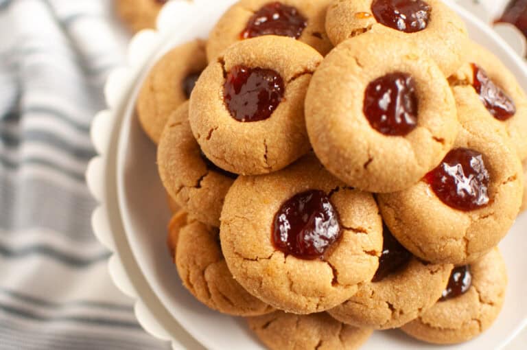 A plate of peanut butter cookies topped with a dollop of red jelly, stacked on a white cake stand.