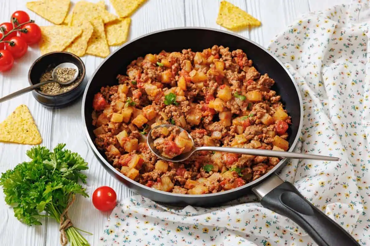 A frying pan filled with Poor Man's meal sits on a white surface with a spoon, surrounded by fresh parsley, tomatoes, tortilla chips, and seasoning.