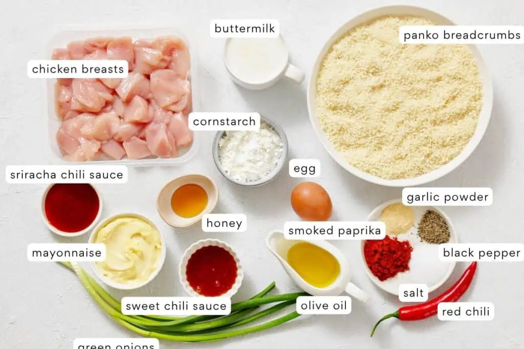 Ingredients for a chicken recipe arranged on a white surface, including chicken breasts, breadcrumbs, sauces, spices, egg, buttermilk, olive oil, cornstarch, mayonnaise, and green onions.