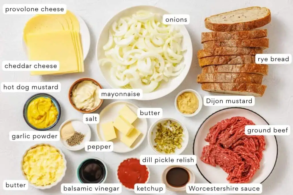 A variety of sandwich ingredients including sliced bread, cheeses, ground beef, sliced onions, condiments, butter, spices, dill pickle relish, and sauces arranged on a white surface.