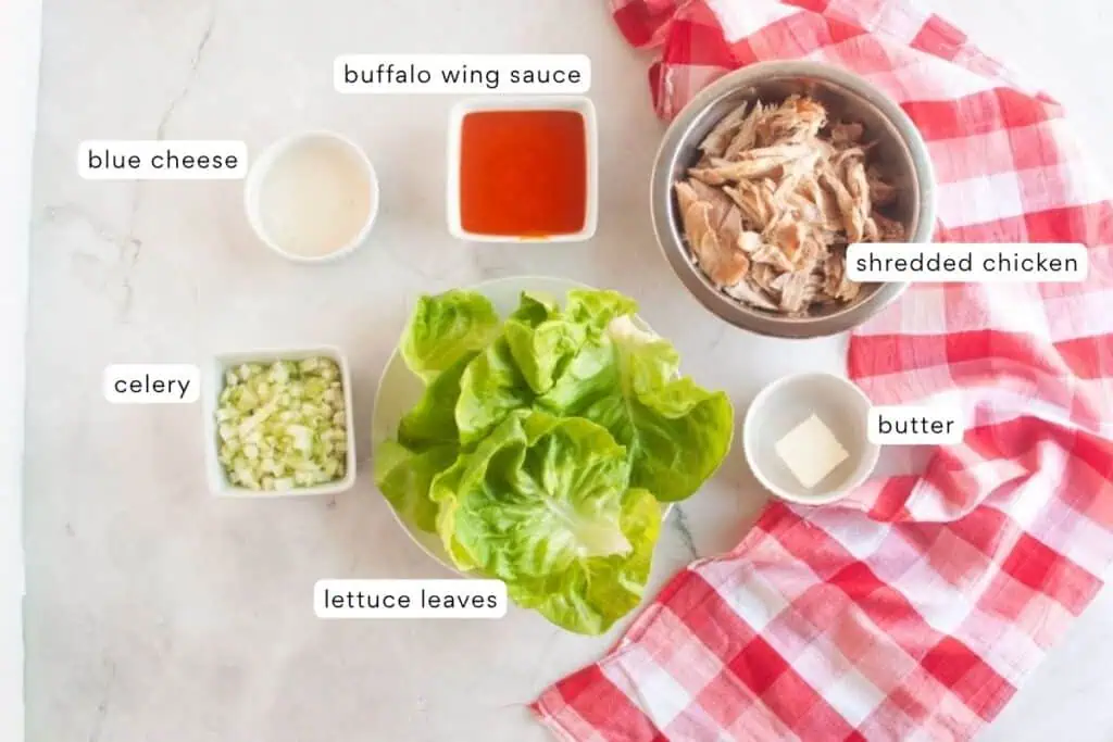 Ingredients for buffalo chicken lettuce wraps arranged on a counter: shredded chicken, buffalo wing sauce, blue cheese, celery, lettuce leaves, and butter, with a red checkered cloth.