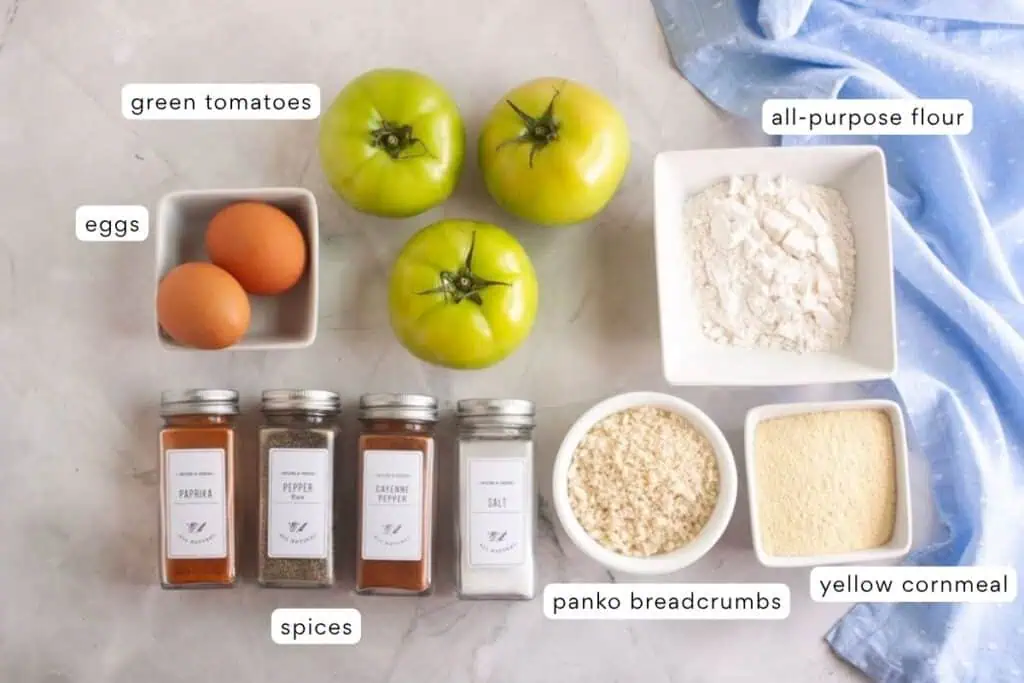 Three green tomatoes, two eggs, all-purpose flour, yellow cornmeal, panko breadcrumbs, and four spice jars are arranged on a countertop with a blue cloth.