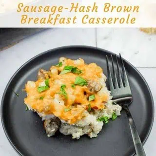 A close-up of a square black plate holding a serving of "Sausage-Hash Brown Breakfast Casserole", featuring a golden-brown melted cheese topping over hash browns and sausage, garnished with fresh chopped parsley, with a black fork on the right.