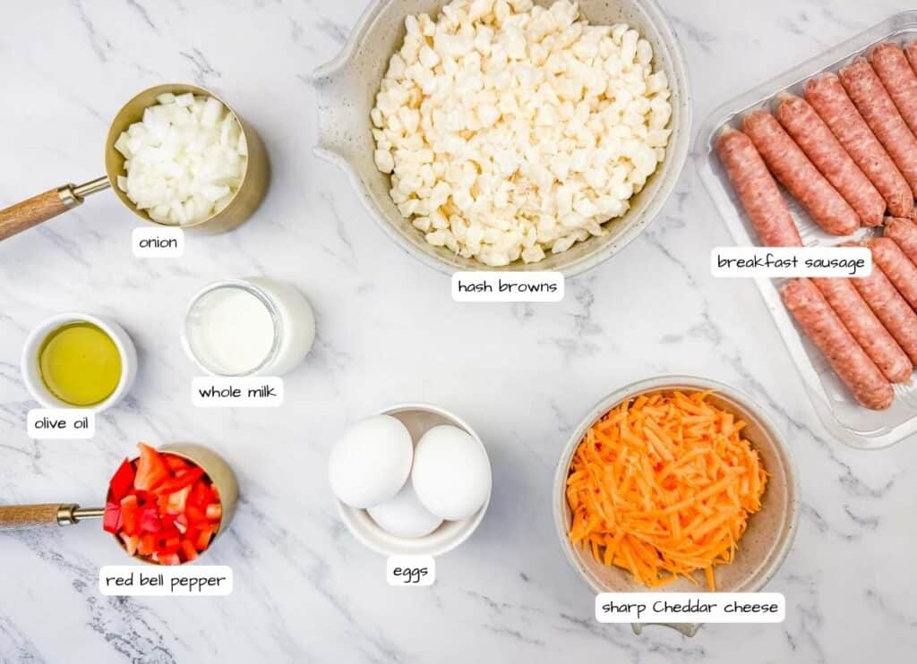 Ingredients for a breakfast casserole on a white surface: diced onion, red bell pepper, olive oil, whole milk, eggs, hash browns, sharp cheddar cheese, and breakfast sausage.