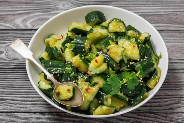 A white bowl filled with chopped cucumber salad garnished with cilantro, sesame seeds, and red pepper flakes, with a spoon resting inside, set on a wooden table.