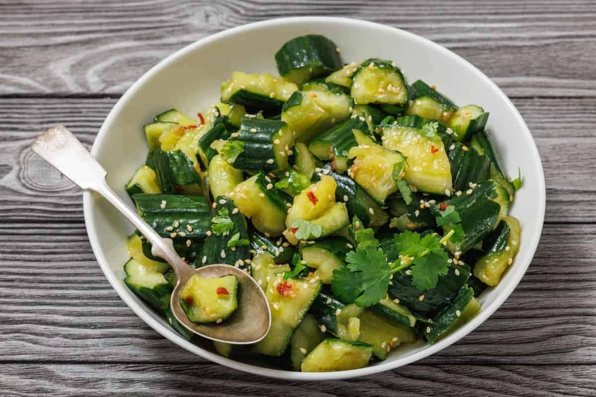 A bowl of smashed cucumber salad garnished with cilantro, sesame seeds, and chili flakes, with a spoon on the side, set on a wooden surface.