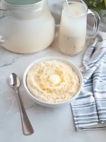 A bowl of creamy grits topped with shredded cheese and a pat of butter sits next to a spoon, a striped napkin, a glass pitcher of milk, and a milk jug on a marble surface.