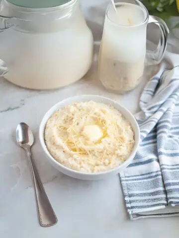 A bowl of creamy grits topped with shredded cheese and a pat of butter sits next to a spoon, a striped napkin, a glass pitcher of milk, and a milk jug on a marble surface.