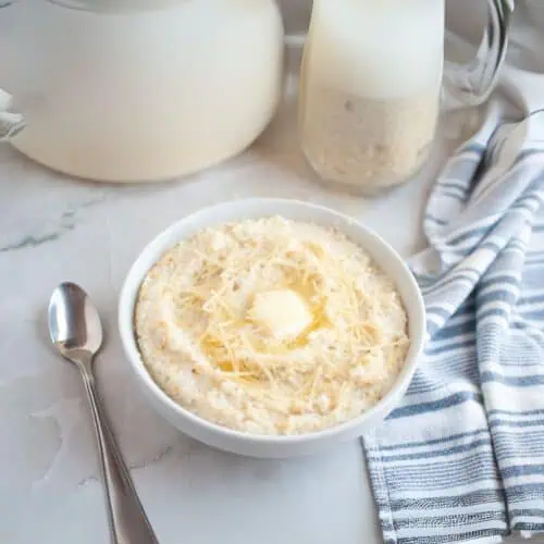 A bowl of creamy grits topped with shredded cheese and a pat of butter sits next to a spoon, a striped napkin, a glass pitcher of milk, and a milk jug on a marble surface.