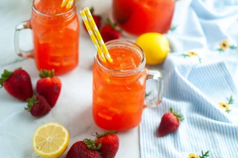 A mason jar filled with iced strawberry lemonade, garnished with two yellow-striped straws, surrounded by fresh strawberries and a halved lemon on a white surface.
