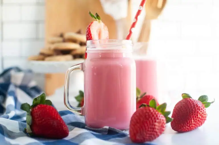 A mason jar filled with pink strawberry milk with a red straw, garnished with a strawberry, surrounded by fresh strawberries on a blue checkered cloth. Cookies are blurred in the background.